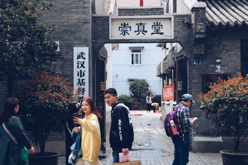 几位年轻人站在一座写着“崇真堂”和“武汉市基督教会”的青砖拱门前。一位穿黄色卫衣的女子正举着手机，画面前景中还能看到一盆红叶植物和提着纸袋的男子。