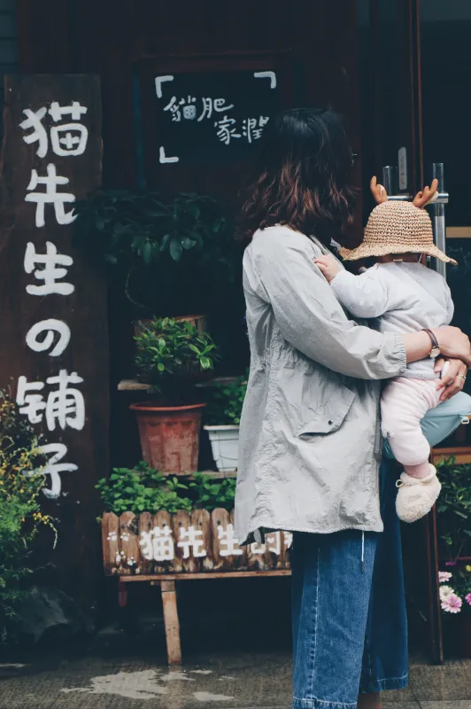 一张站在一家店铺门口的女性背影照片，女性身穿灰白色风衣抱着一个戴草帽的婴儿。