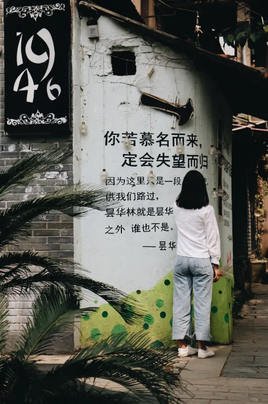 一名穿白色上衣和浅色牛仔裤的女子背对镜头，站在一面写着文字的灰墙前观看。女子身旁有绿色的植物叶片，墙上挂着复古喇叭和黑白招牌。