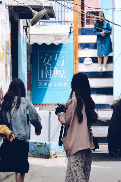 三名身穿便装的女性正在一条狭窄的街道中行走，前方墙壁上有一处蓝色的'安与南鲸'招牌，右侧有通往高处的楼梯。