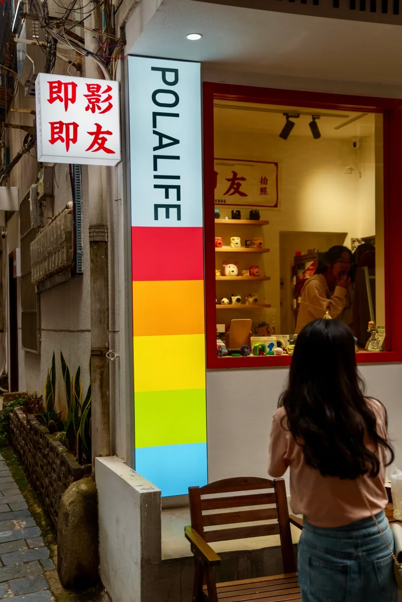 一位长黑发女子背对镜头站在红框窗口外，窗内一位穿米色卫衣的人低头整理，旁边立着写有 POLALIFE 与“即影即友”的彩色条纹招牌，木椅置于墙边。