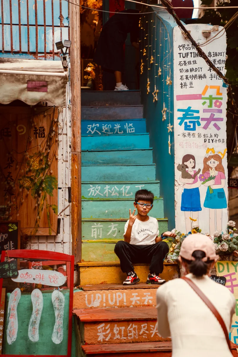 一个戴眼镜的小男孩坐在彩色的户外楼梯台阶上，面对镜头比出剪刀手。前景中一位戴着帽子的女士背对镜头正在为他拍照，背景是色彩斑斓的店铺入口和手绘涂鸦。