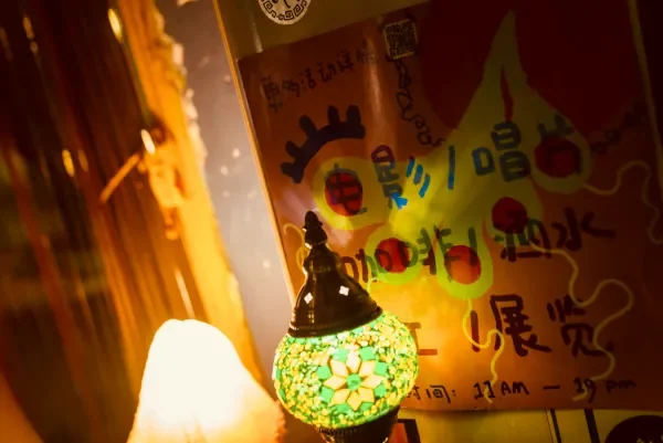 画面前景是一个发光的绿黑马赛克灯罩，背景是一张手写海报与模糊的室内角落。