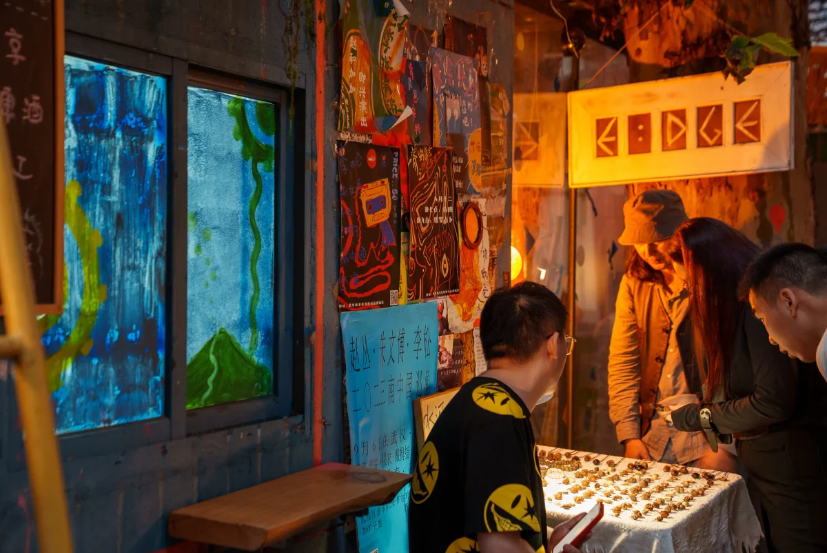 画面展示了一个夜晚的小摊位场景。左侧是绘有蓝色与绿色抽象图案的窗户墙壁，墙边张贴着海报。中间一位戴眼镜的男子正在低头看手机，右侧桌面上铺着白色桌布，摆放着许多小件工艺品。一名戴着帽子的摊主与一位长发顾客正站在桌前挑选商品，旁边还有一位短发男子在围观。