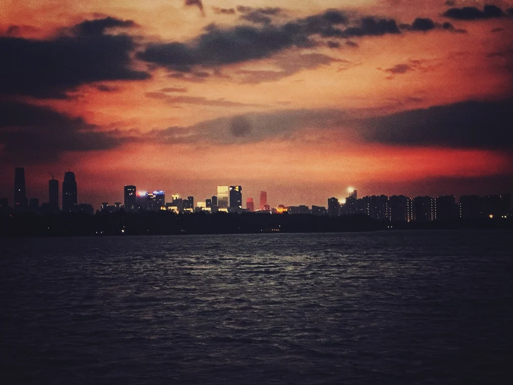 画面中是一片广阔的水域，远处是城市天际线的黑色剪影，天空被浓烈的橙红色云层覆盖，城市中亮起的灯光点缀在暗色轮廓上。